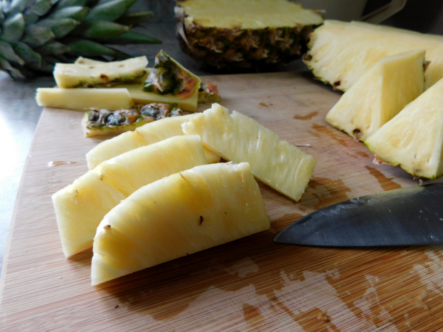 Close-up of cut pineapple chunks on a wooden cutting board, the tip of heavy knife visible in the bottom right corner.