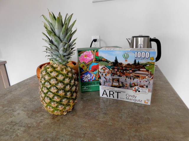 A kitchen table with an electric kettle just visible in the background. A large fresh pineapple sits in the foreground beside two puzzles boxes. 