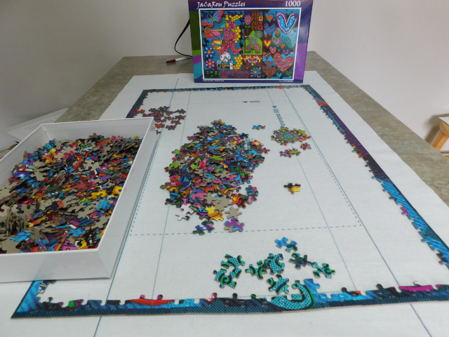 A brightly colored jigsaw puzzle is in its early stage of being put together. The outside pieces are in place; there's a heap of pieces in the middle of the puzzle mat and even more pieces in the box at the side. The box cover with the picture of many brightly colored hearts sits upright in the background.