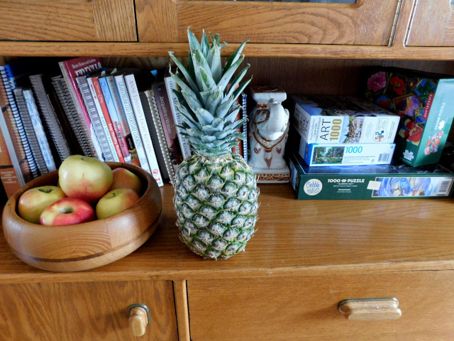 A whole pineapple in the foreground beside a wooden bowl of apples, on the  shelf of an oak china cabinet. In the background are several recipe books, a china elephant serving as book end and a few puzzles. 