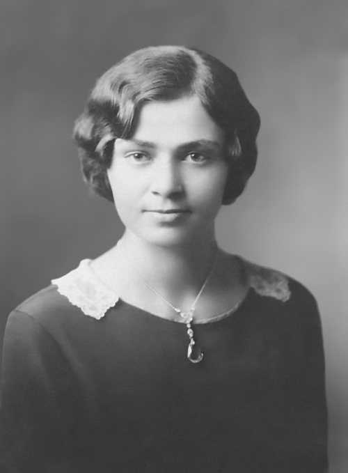 Photo of my mother. Her hair is dark, carefully waved back from her face in the style of the early 1930s. She wears a simple dark dress with a small lace color and a pendant. Her eyes have a clear gaze and her mouth looks as if she might smile but she doesn't quite smile. Her skin appears flawless. 